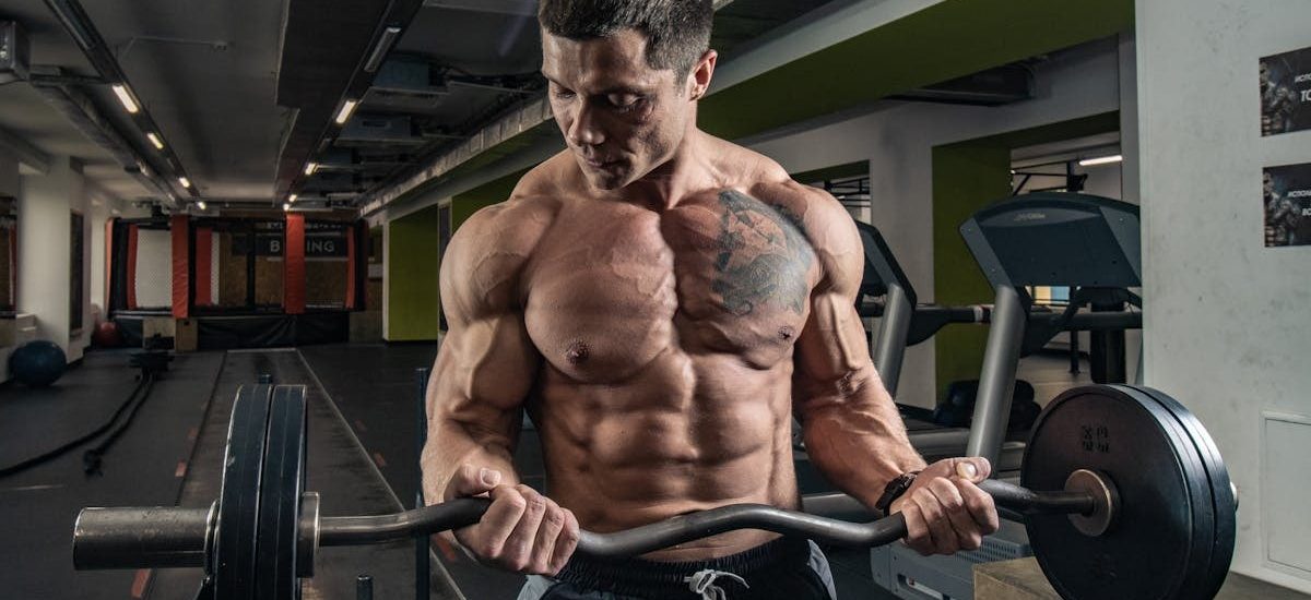Topless bodybuilder with tattoos lifting weights in an indoor gym setting.