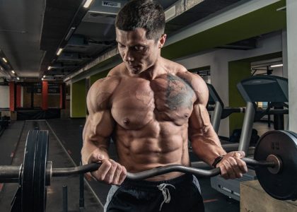 Topless bodybuilder with tattoos lifting weights in an indoor gym setting.